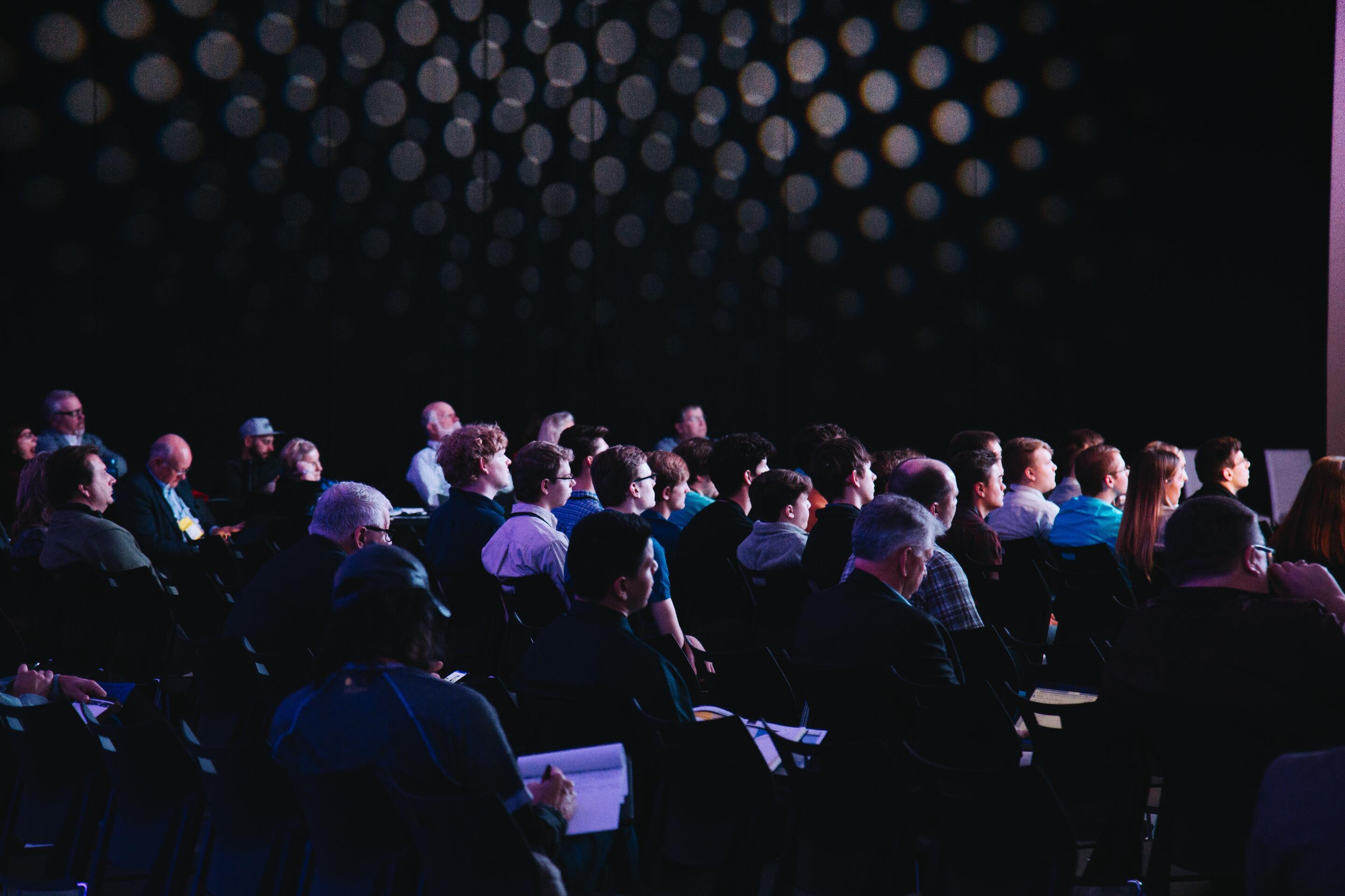 An audience sits attentively in a dark room, listening to a presentation or lecture. The room is decorated with circular light patterns, creating a unique atmosphere. The people are of various ages and genders, all focused on the front.