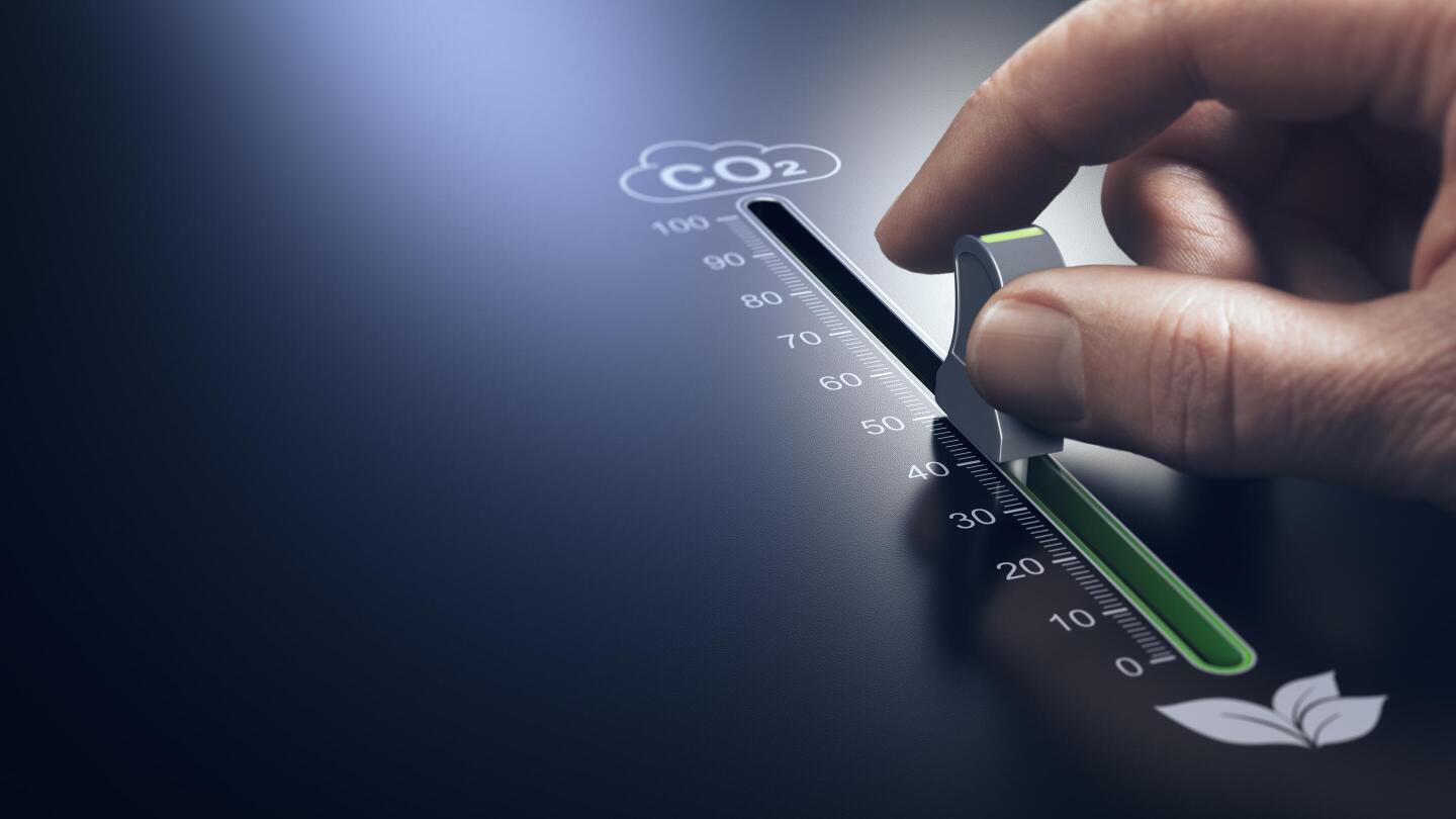 A hand moves a slider on a CO₂ scale from a high value towards zero, symbolizing the reduction of emissions.