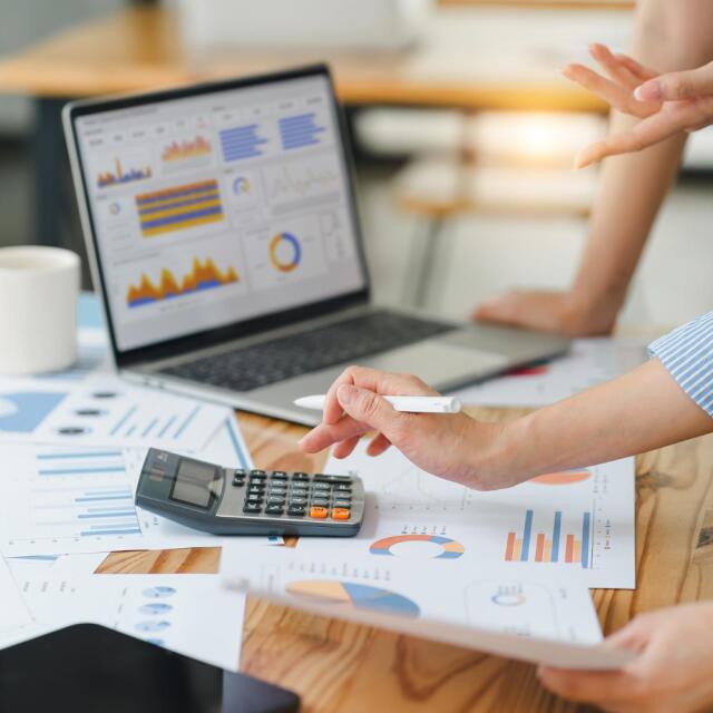 Nahaufnahme von Händen, die einen Taschenrechner auf einem Schreibtisch voller Finanzdiagramme und einem Laptop mit Datenvisualisierungen nutzen.