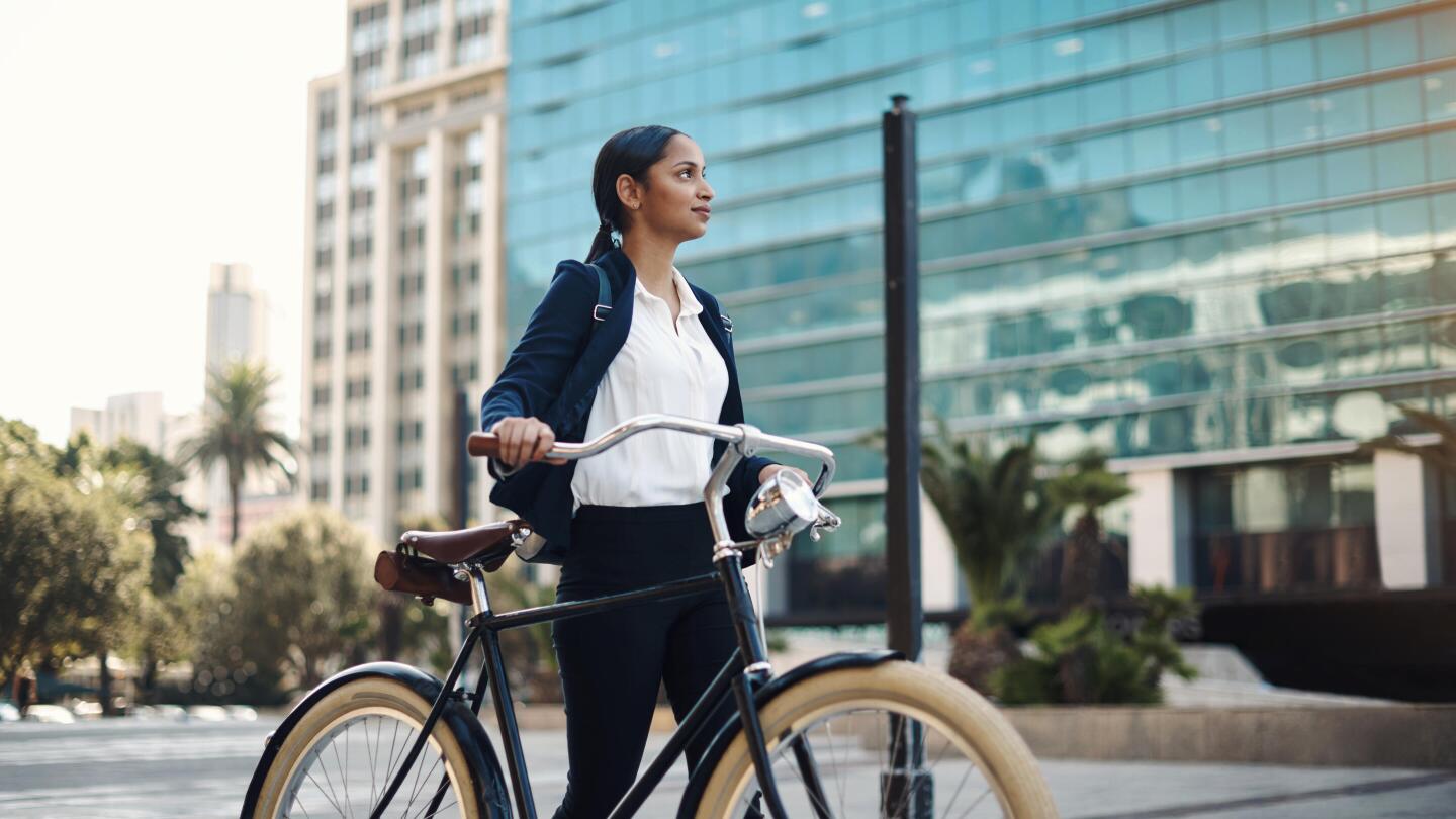 A businesswoman in a suit stands with her bicycle in a modern city with glass buildings.