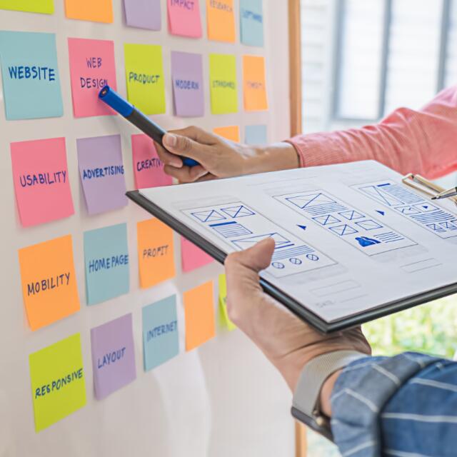 Marketing-Team stimmt Aufgaben an einem Whiteboard mit Post-its und Entwürfen für ein strukturiertes Projekt-Management ab.