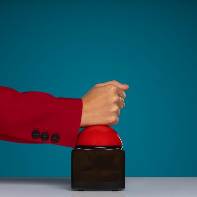 Hand im roten Blazer drückt auf einen großen roten Buzzer-Knopf vor türkisem Hintergrund.