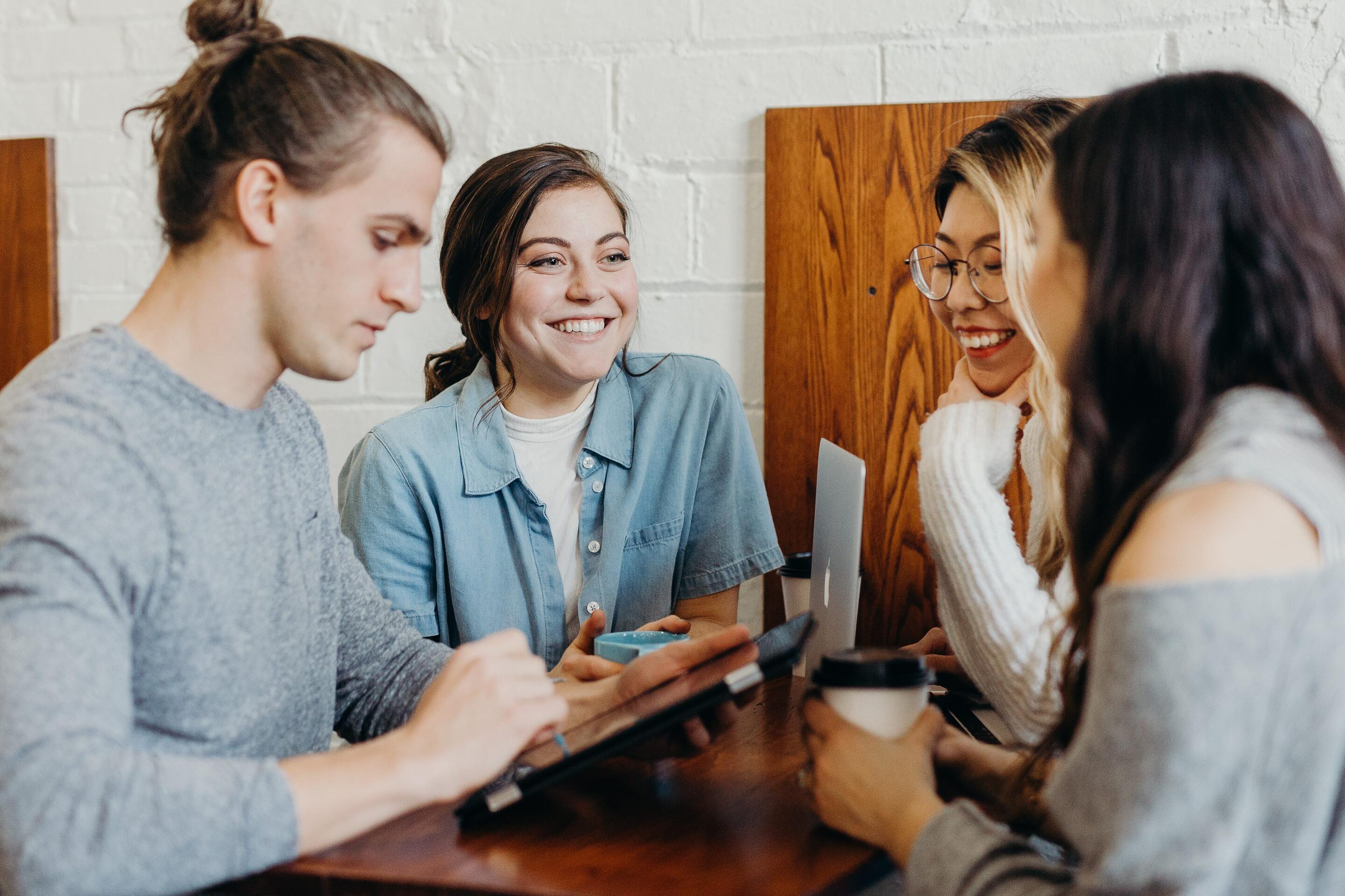 Four young adults sit at a wooden table in a cozy café or workspace. They are having a cheerful conversation, laughing, and collaborating using a tablet and laptop. The atmosphere is relaxed and creative.