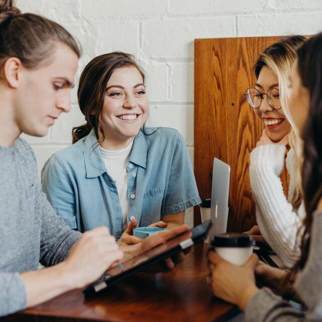 Vier junge Erwachsene sitzen an einem Holztisch in einem gemütlichen Café oder Arbeitsraum. Sie unterhalten sich fröhlich, lachen und arbeiten gemeinsam an einem Tablet und Laptop. Eine entspannte und kreative Atmosphäre ist spürbar.