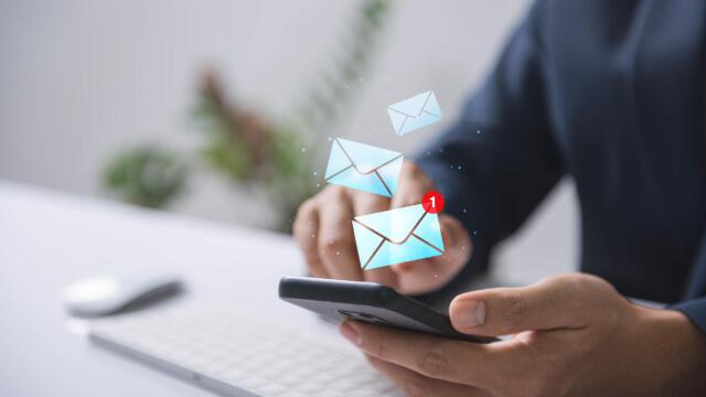 Person hält ein Smartphone in der Hand, auf dem eine neue E-Mail-Benachrichtigung angezeigt wird; im Hintergrund ist eine Computertastatur zu sehen.