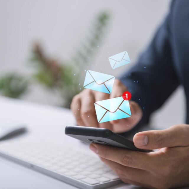 Person hält ein Smartphone in der Hand, auf dem eine neue E-Mail-Benachrichtigung angezeigt wird; im Hintergrund ist eine Computertastatur zu sehen.
