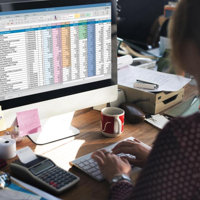 Eine Person arbeitet an einem Schreibtisch an einer “Marketing Budget”-Tabelle auf einem großen Computermonitor, umgeben von Büromaterial.
