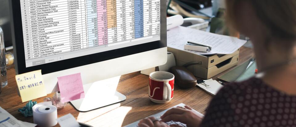 A person works at a desk on a “Marketing Budget” spreadsheet displayed on a large computer monitor, surrounded by office supplies.