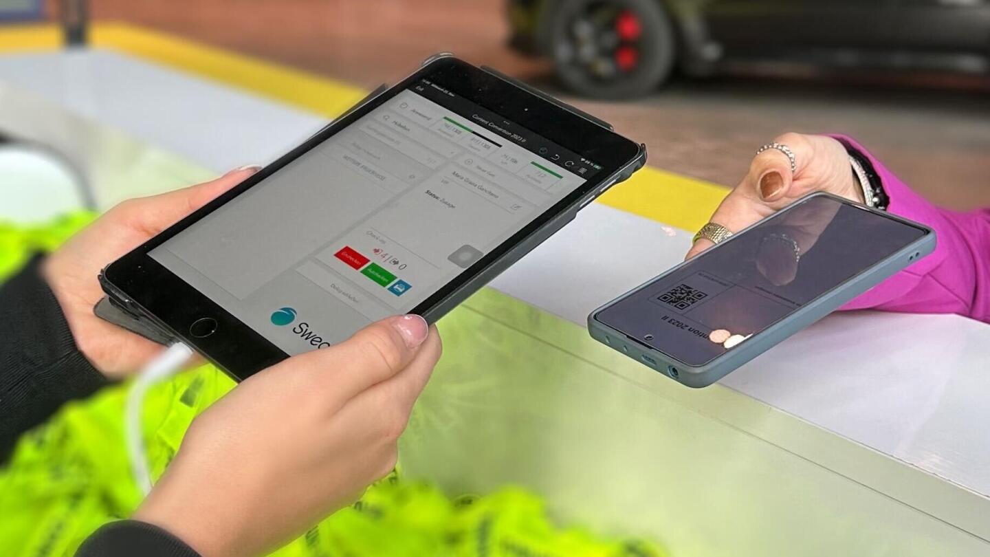 A person holds a smartphone with a QR code up to a tablet at an event check-in, which is running the Sweap software.