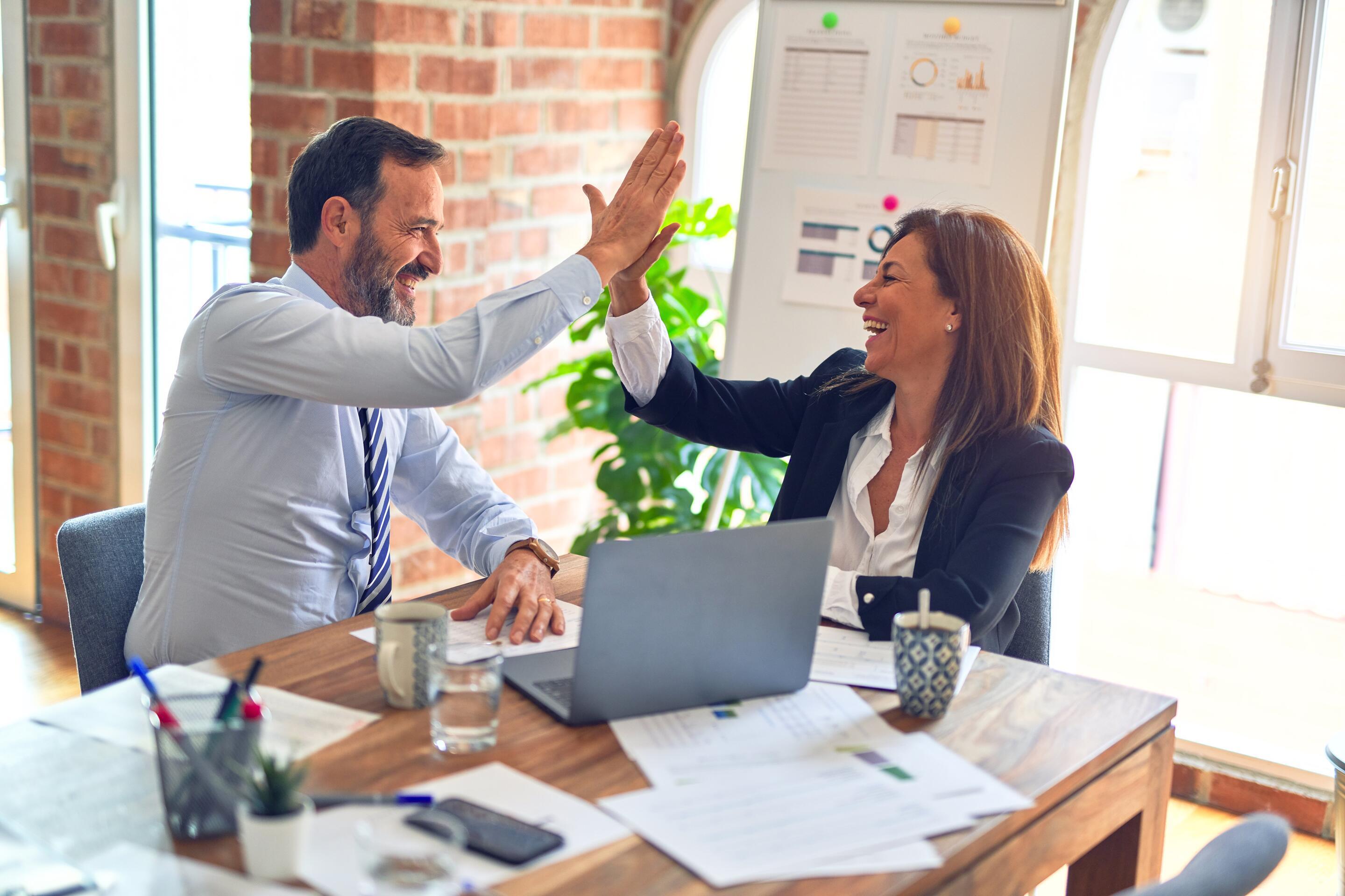 Two colleagues in an office share a cheerful high-five at a desk with a laptop and documents.