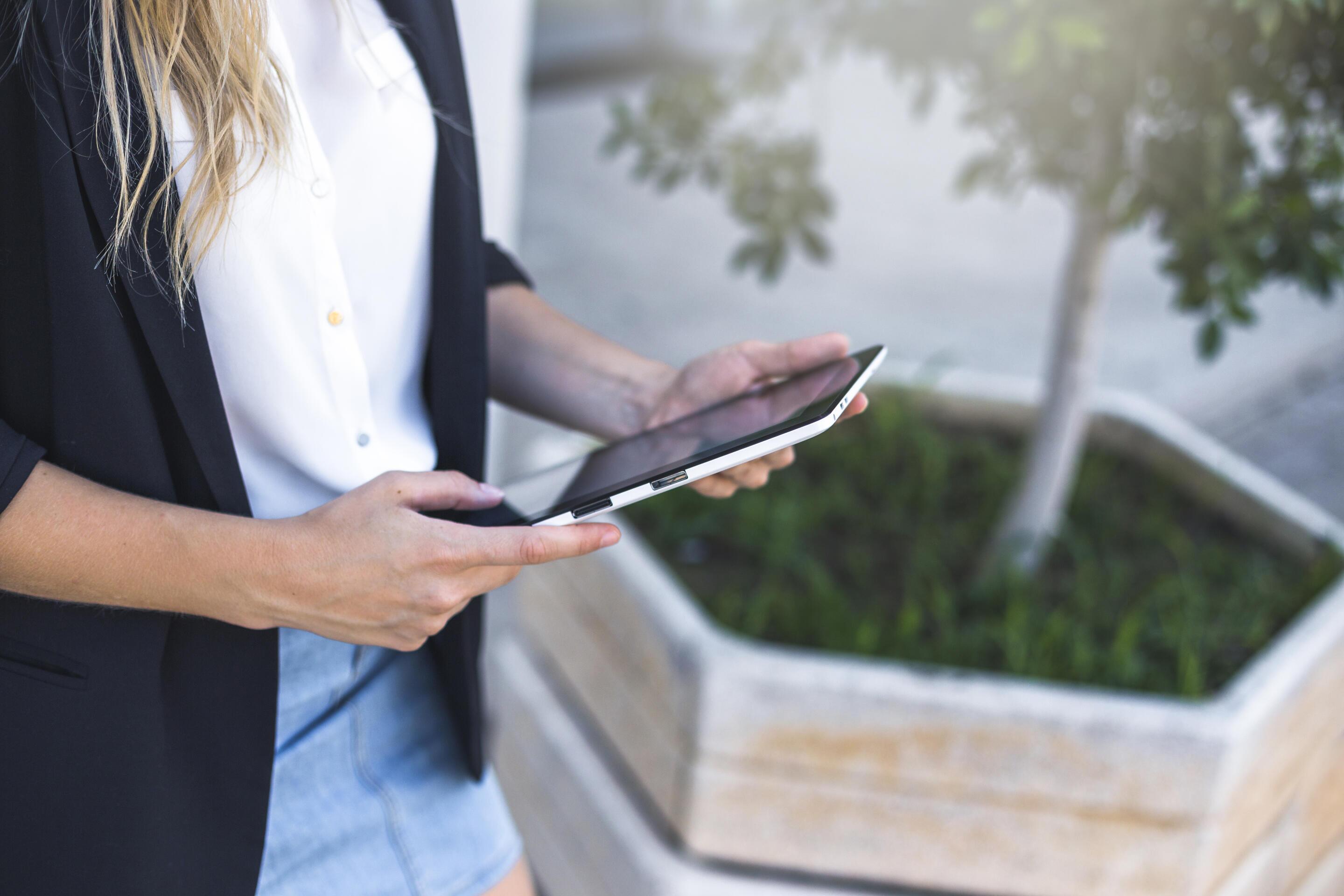 Woman in business attire holding a tablet outdoors next to a large planter.