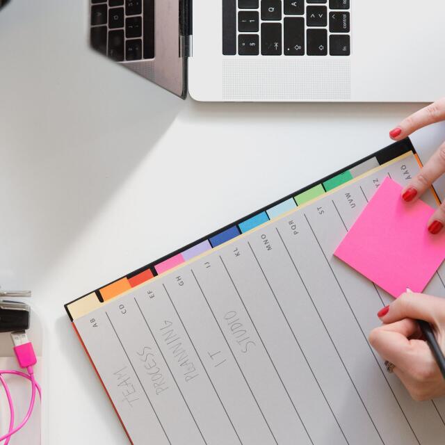 Eine Person sitzt an einem Schreibtisch mit einem Laptop, schreibt auf einen pinkfarbenen Notizzettel und hat eine Planungsmappe mit verschiedenen farbigen Registerkarten vor sich. Auf dem Tisch liegen außerdem Büroartikel wie ein Textmarker, Kopfhörer und ein Stift.