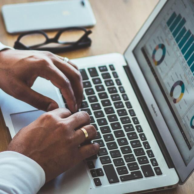 Eine Person arbeitet an einem Laptop, auf dessen Bildschirm Diagramme und Balkengrafiken zu sehen sind. Auf dem Tisch liegen außerdem eine Brille und ein Smartphone.