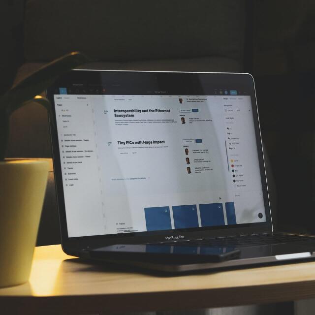 Ein Laptop steht auf einem kleinen Tisch neben einer Pflanze. Auf dem Bildschirm sind Webseiten oder Online-Dokumente mit Texten und Profilbildern zu sehen. Vor dem Laptop liegt ein Smartphone.