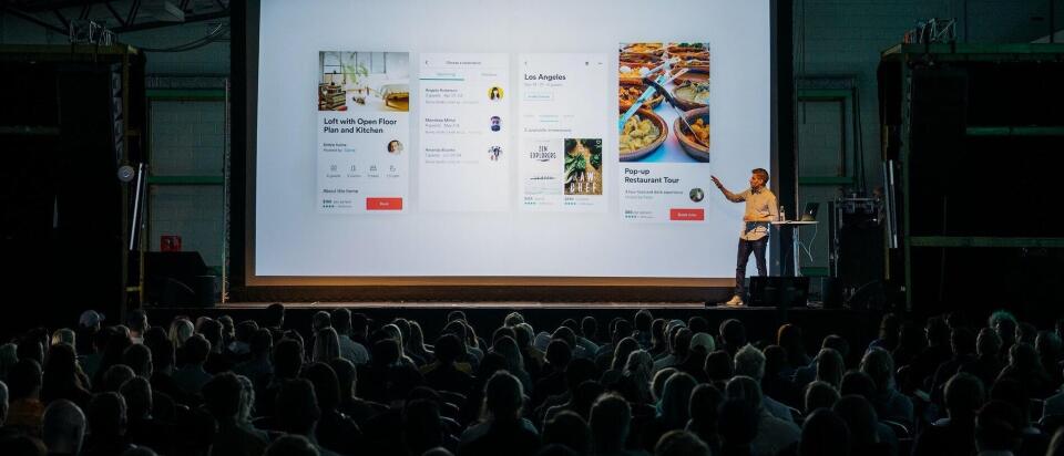 Ein Sprecher steht auf einer Bühne und präsentiert vor einem großen Publikum eine Bildschirmpräsentation mit verschiedenen App- oder Website-Designs, die auf einer großen Leinwand angezeigt werden.