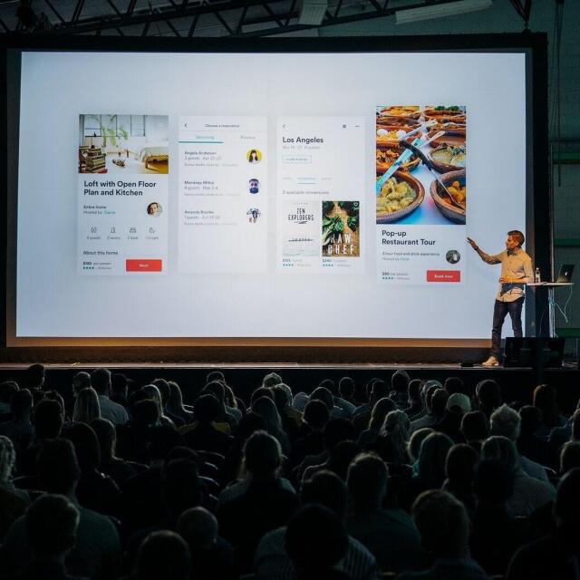 Ein Sprecher steht auf einer Bühne und präsentiert vor einem großen Publikum eine Bildschirmpräsentation mit verschiedenen App- oder Website-Designs, die auf einer großen Leinwand angezeigt werden.