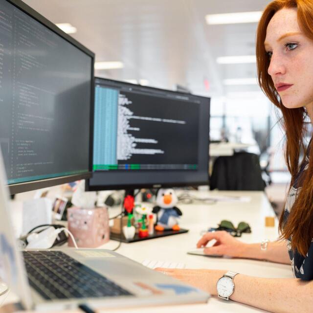 Eine Frau arbeitet an einem Schreibtisch mit mehreren Monitoren, auf denen Programmiercode angezeigt wird. Sie konzentriert sich auf ihre Arbeit in einem modernen Büro.