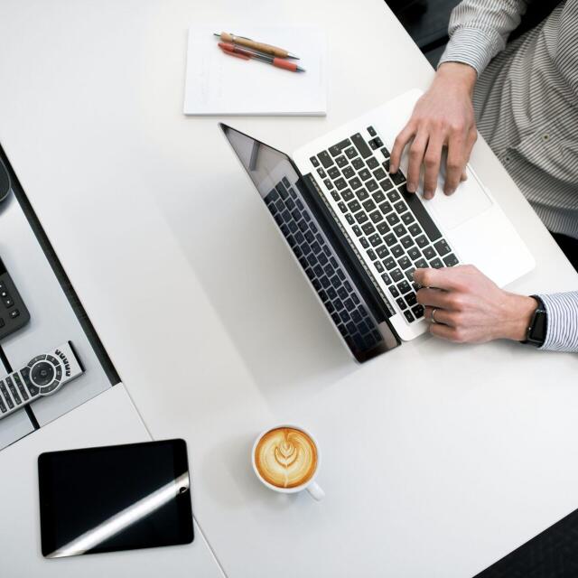 Ein Mann arbeitet an einem Laptop an einem modernen Schreibtisch. Neben ihm stehen eine Tasse Kaffee, ein Tablet, ein Telefon und einige Stifte auf einem Notizblock.