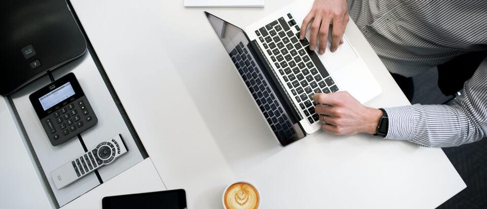 Ein Mann arbeitet an einem Laptop an einem modernen Schreibtisch. Neben ihm stehen eine Tasse Kaffee, ein Tablet, ein Telefon und einige Stifte auf einem Notizblock.